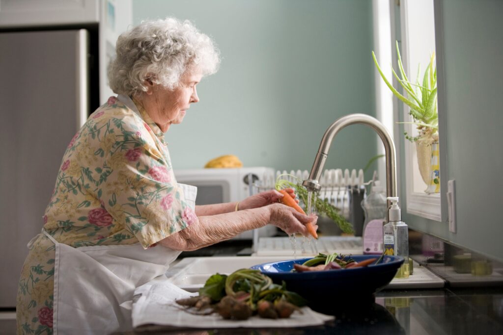 Older woman at kitchen sink with apron on, washing a carrot getting ready to prep a meal