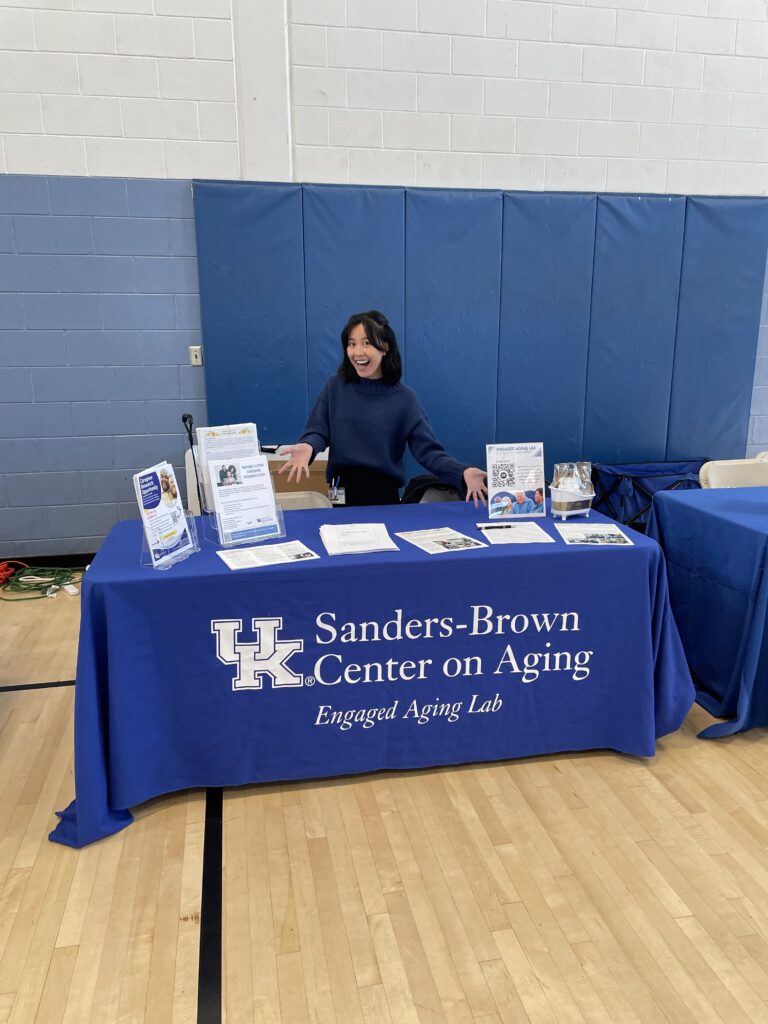 Nancy stands behind Engaged Aging Lab table gesturing towards health resource flyers on table